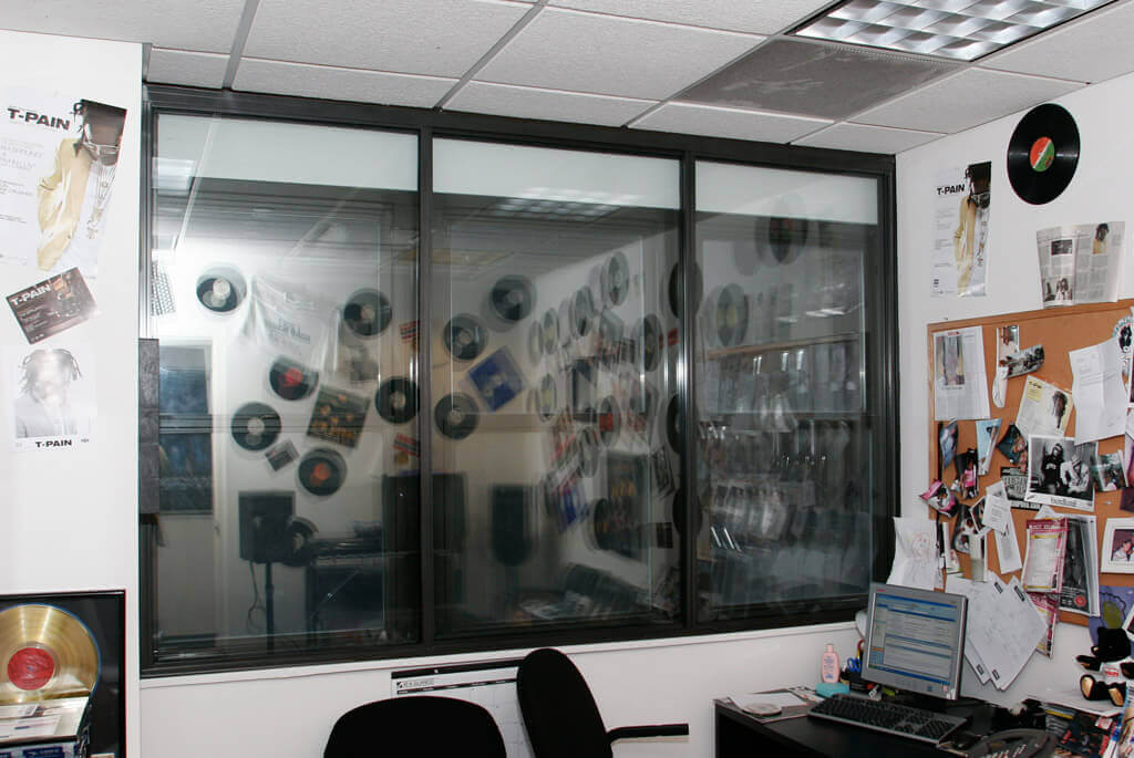 Office with a glass window displaying vinyl records on the other side, a corkboard with papers and photos, a computer on a cluttered desk, and framed awards on the walls. Two chairs are positioned by the desk.