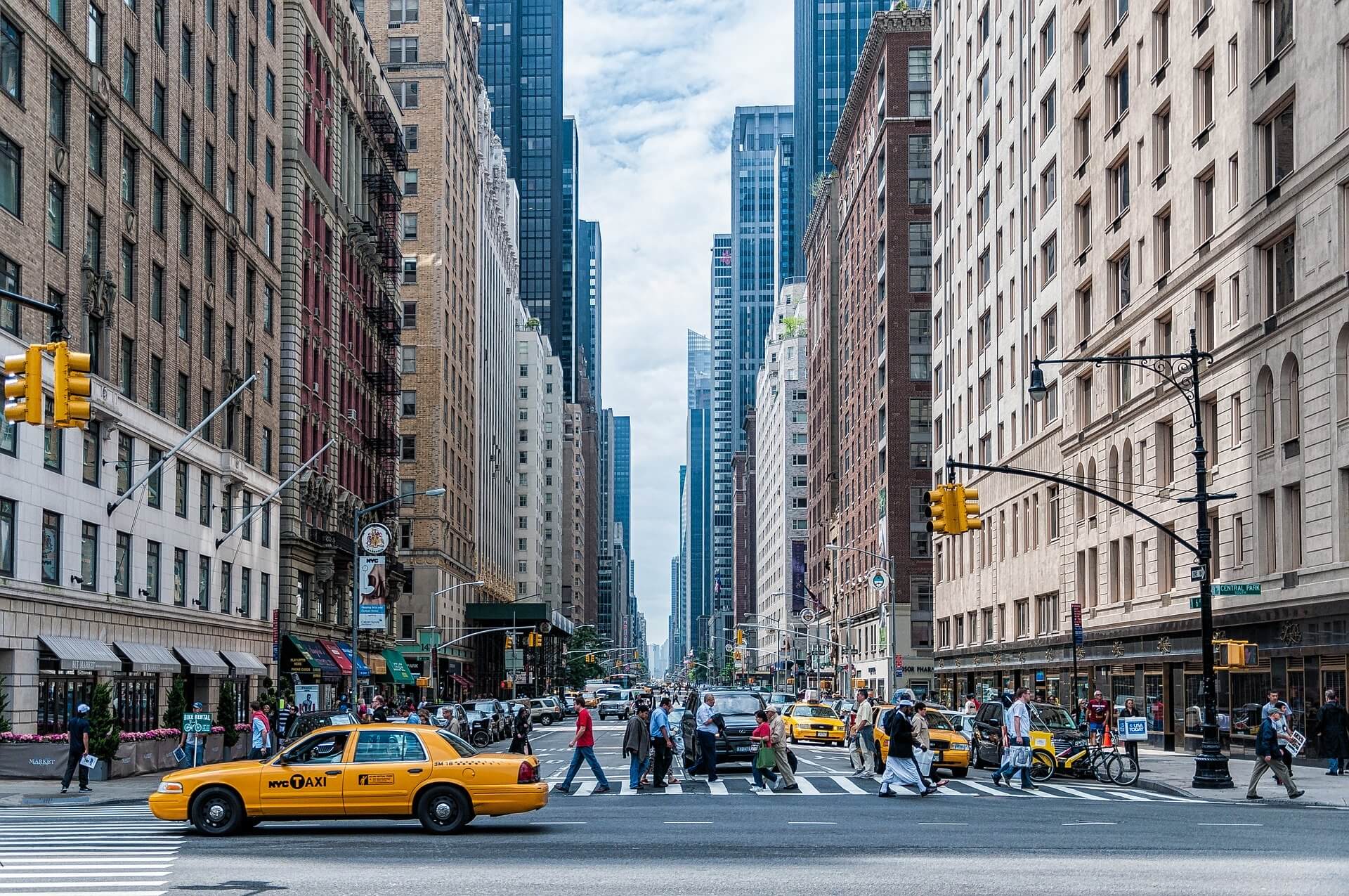 City street scene with tall buildings on both sides, several yellow taxis, and people crossing a busy intersection on a cloudy day. Traffic lights and street signs are visible along the sidewalks.