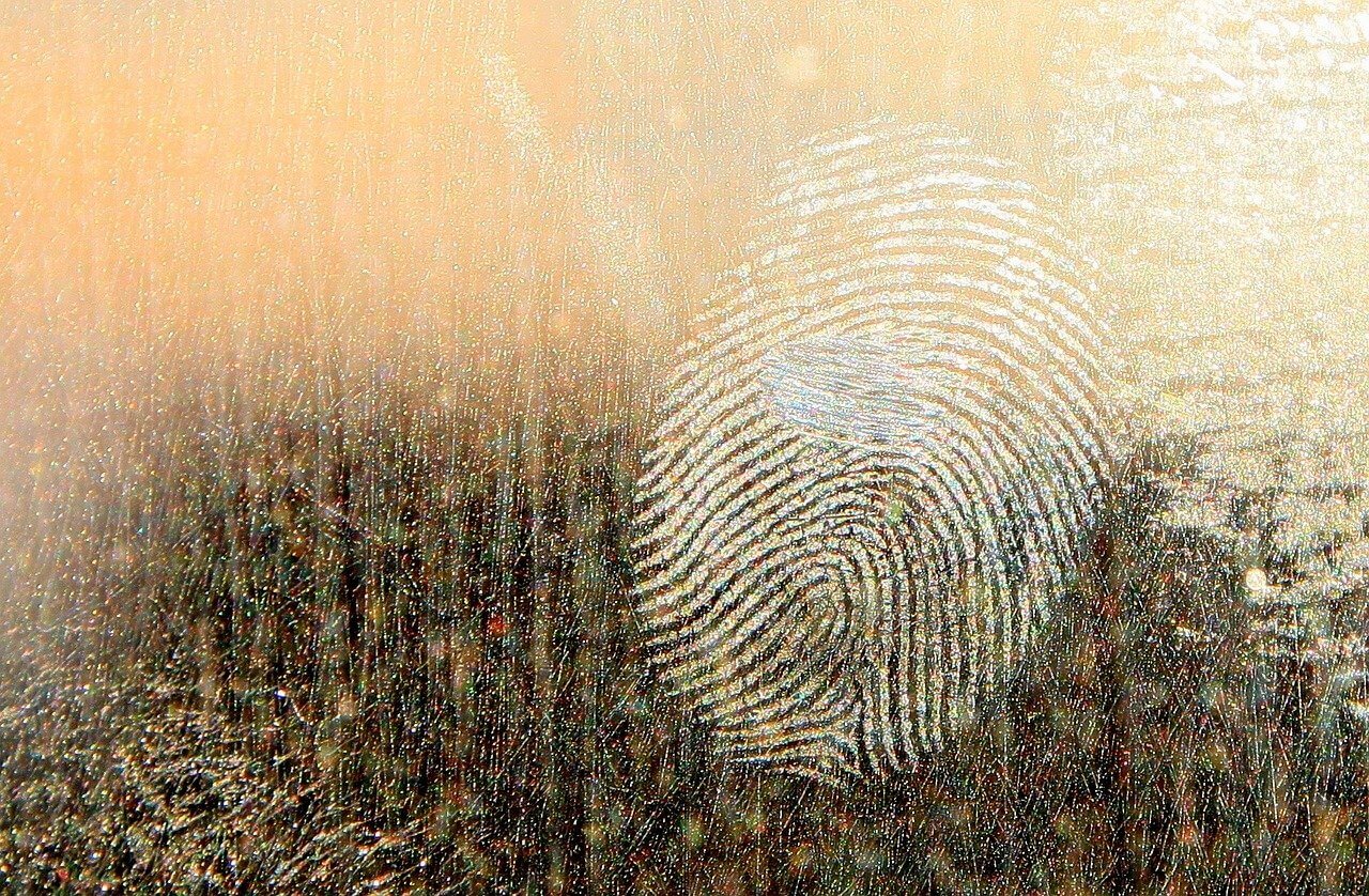 A close-up of a visible fingerprint on a smooth, reflective surface. The fingerprint ridges are detailed and contrast with the textured, slightly shimmering background.
