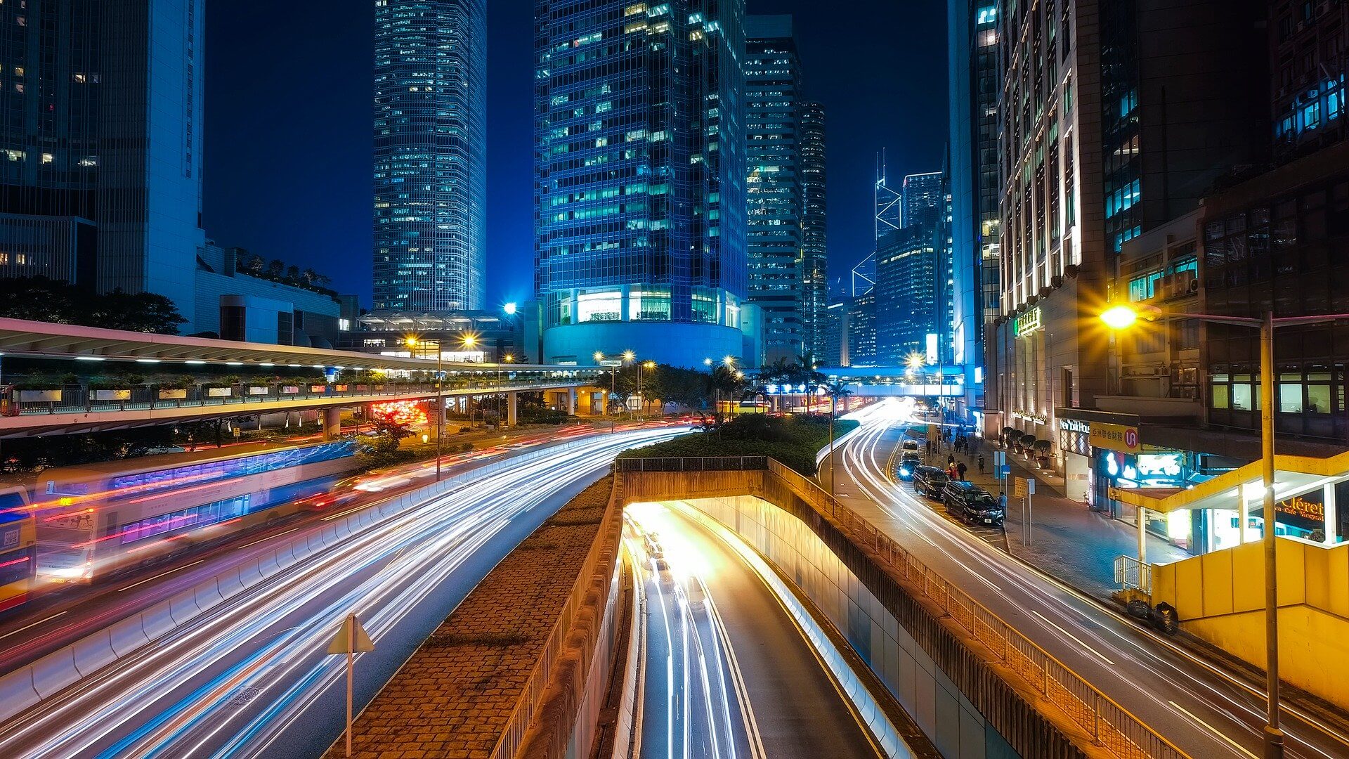 A cityscape at night with tall skyscrapers, illuminated office windows, and busy streets. Light trails from vehicles indicate fast-moving traffic on roads and highways, with streetlights and signs visible.