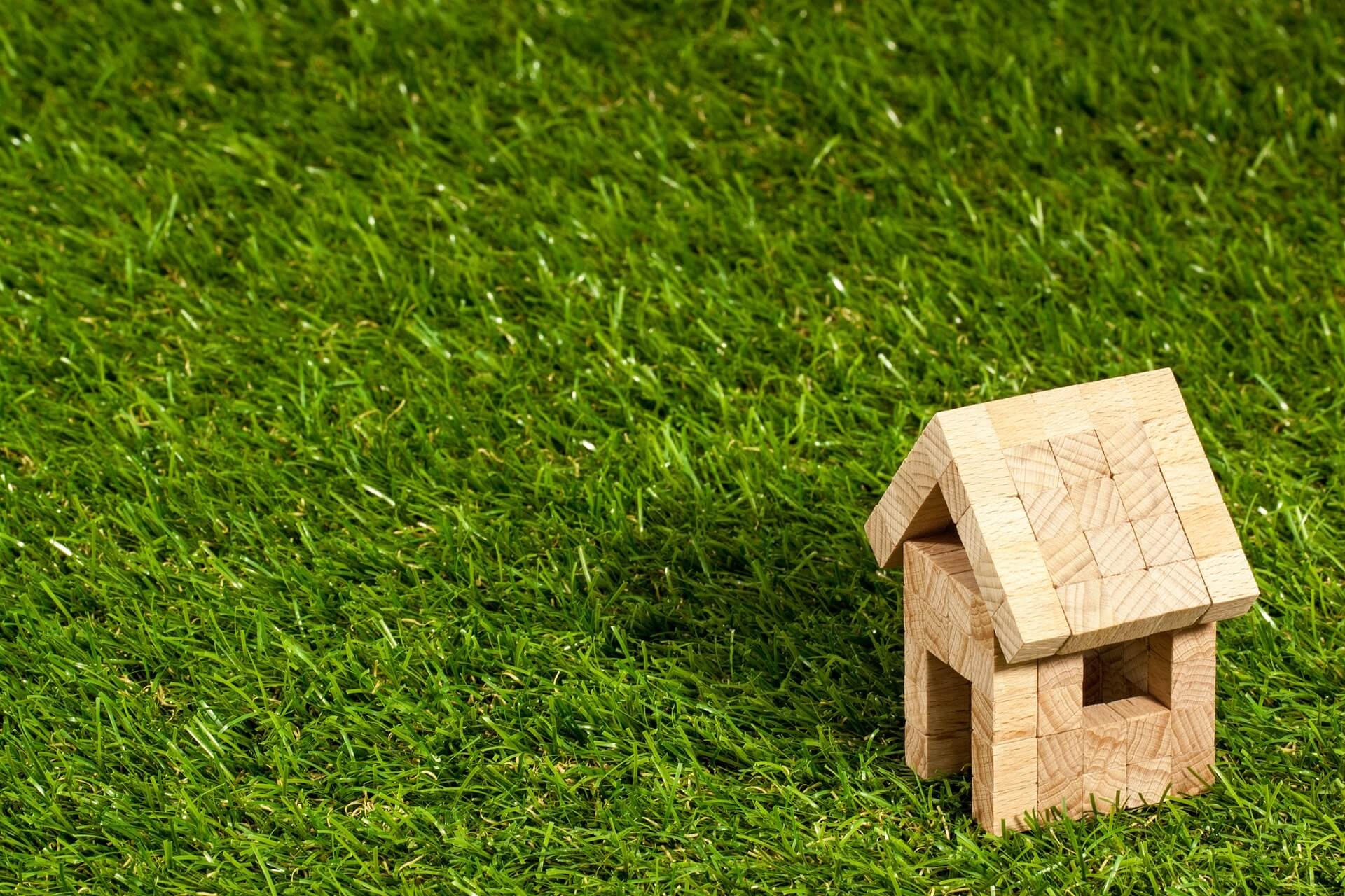 A small wooden house model sits on green grass, positioned in the lower right corner of the image, with the rest of the space showing empty grass.