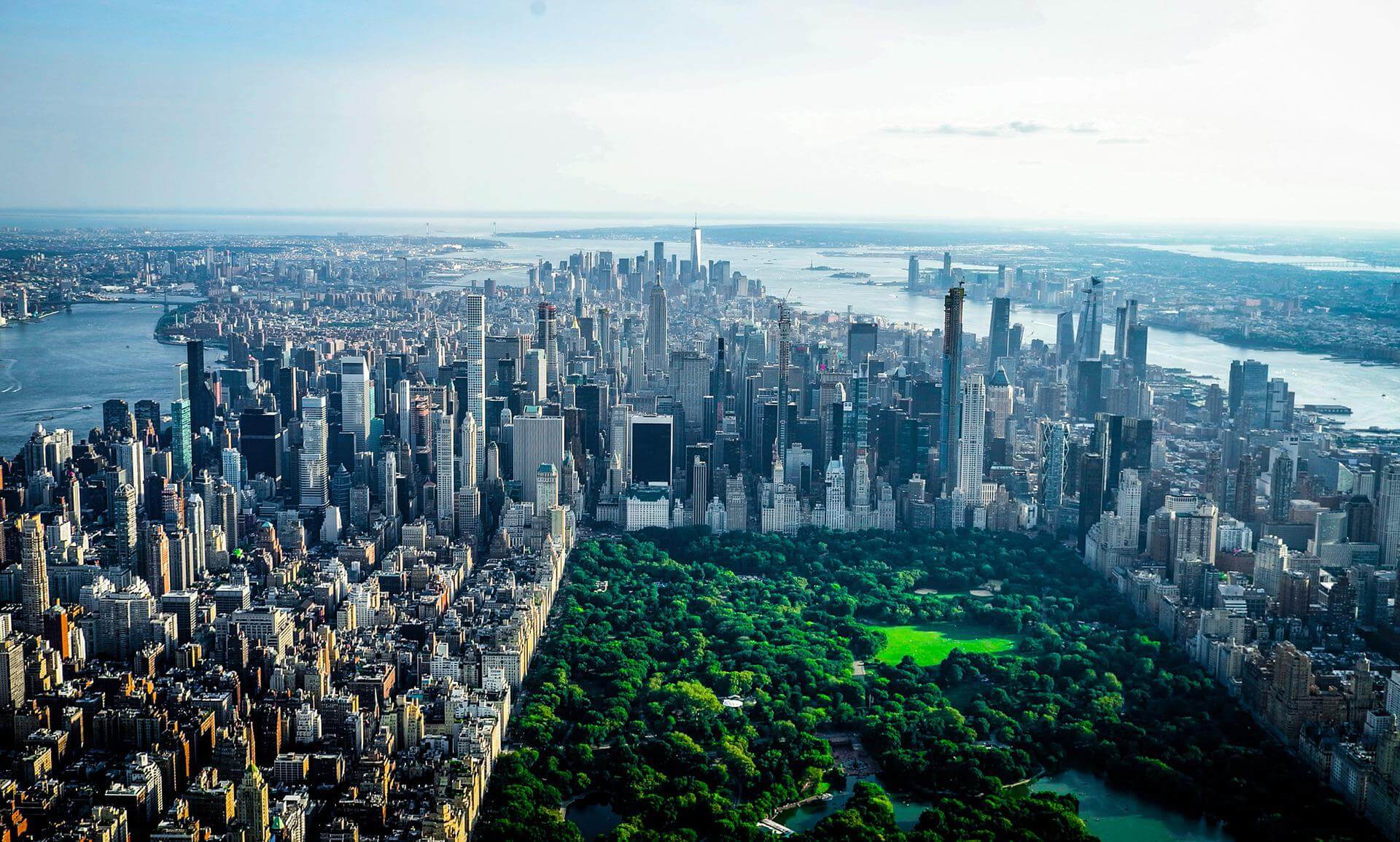 Aerial view of Central Park in New York City, surrounded by tall skyscrapers and dense urban buildings, with rivers visible on both sides of Manhattan.