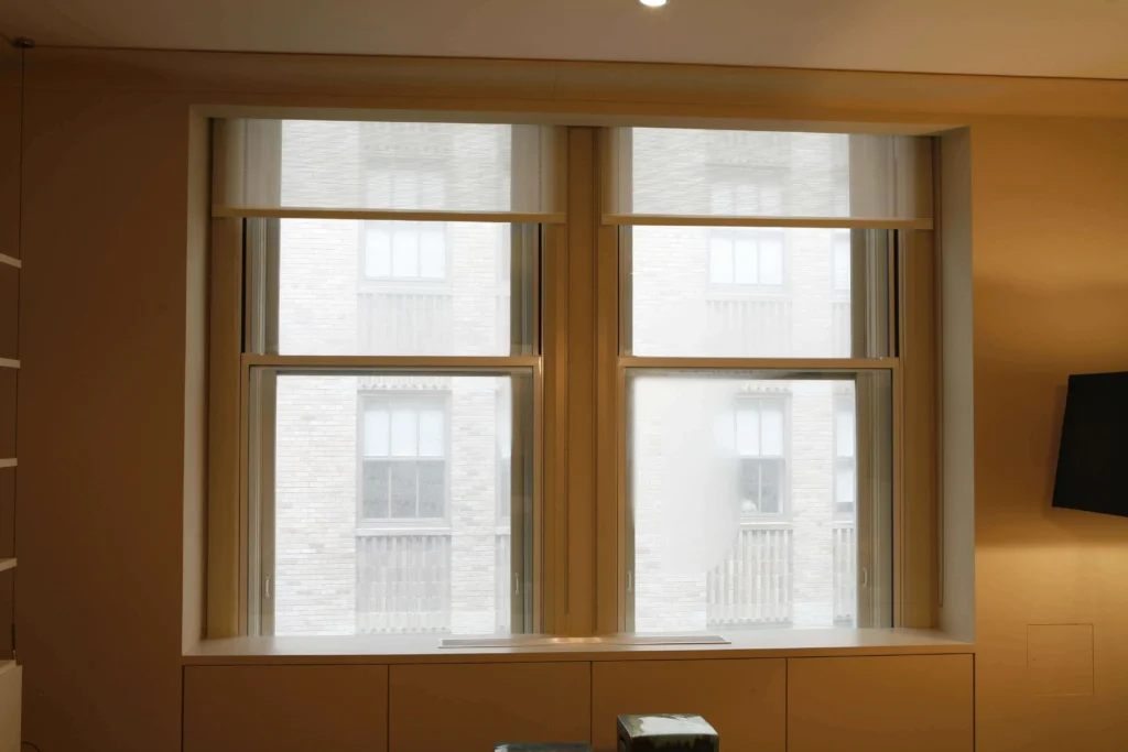Two large windows with white frames are set in a beige wall. Outside, a brick building with multiple windows is visible. The room’s interior includes a black television mounted on the right and a white shelf on the left.