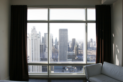 A city skyline with tall buildings is visible through a large window with dark curtains. Part of a white sofa is visible in the lower right corner of the room.