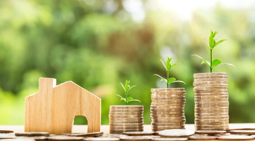A wooden house figure sits next to three stacks of coins, each stack topped with a small plant sprout. The background is green and sunlit, suggesting growth and financial investment in real estate.