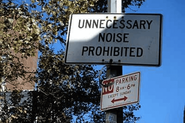 A street pole with two signs: the top sign reads "UNNECESSARY NOISE PROHIBITED" and the lower sign states "NO PARKING 8AM-6PM EXCEPT SUNDAY." Tree branches and a building are visible in the background.