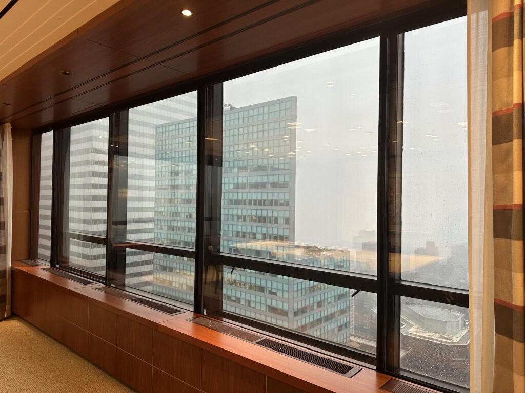 Large floor-to-ceiling windows offer a view of tall office buildings on a foggy or overcast day. The interior has wood paneling and a carpeted floor.