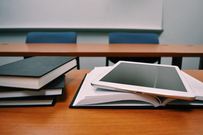 Tablet resting on an open notebook beside stacked books on a wooden desk in a meeting room.